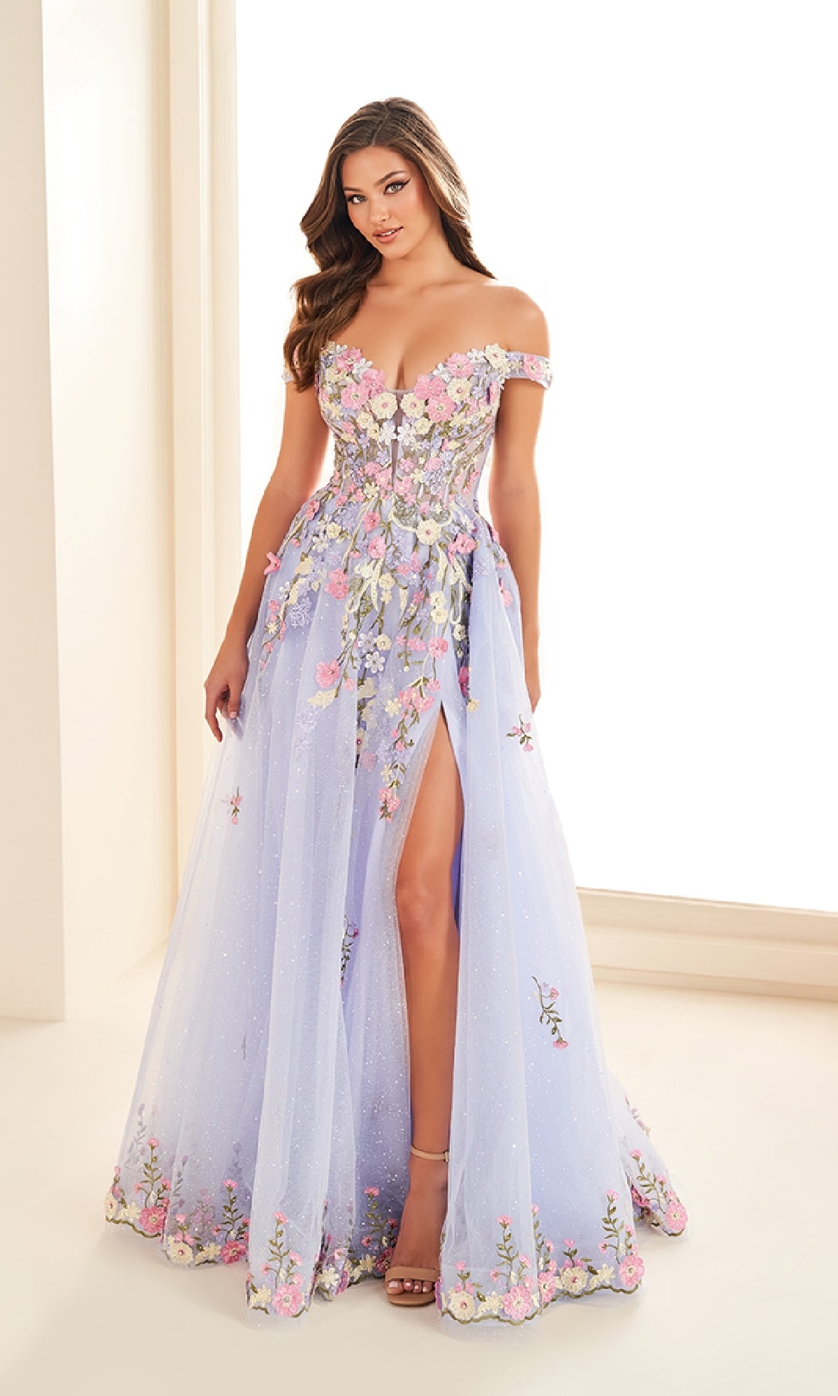 A woman models the Ellie Wilde EW36215 long prom dress—light purple, off-the-shoulder, with floral appliqués and a high leg slit—standing indoors against a light background.